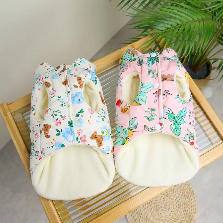 Two dog coats, one white with floral and teddy bear print, and the other pink with floral print, displayed on a wooden tray.